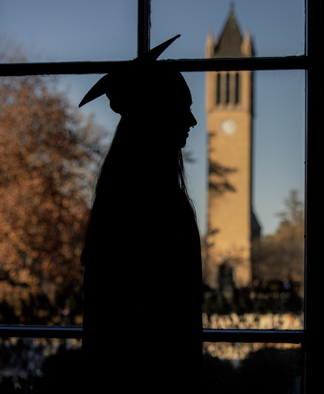 Silhouette of a female student in graduation regalia, standing in front of a window with the ISU Campanile visible outside.